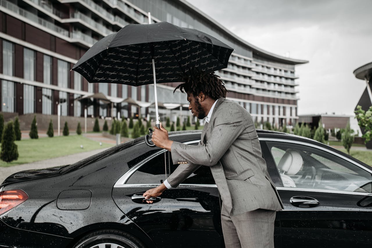 African American man in suit with umbrella opening car door outside modern building.